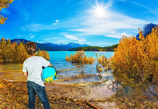 Exquisite Abraham Lake with turquoise water. Boy in jeans with a globe in his hands admires the lake. Indian Summer in the Rockies. Canada. Concept of ecological and active tourism