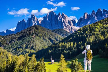 Etkin yaşlı kadın turist sırt çantası ile kilise Santa Maddalena Dolomites içinde fotoğraflar. Ormanlık dağlar Alpin çayırlar tarafından çevrili. Ekolojik ve aktif Turizm kavramı