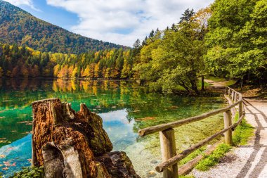 Lago de Fusine gölünün etrafındaki yol ahşap korkuluklarla çevrili. Ekolojik ve kültürel turizm kavramı. Yeşil suyun pürüzsüz yüzeyi bulutlu gökyüzünü yansıtır.
