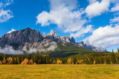 Kanada Kayalıkları 'nda verimli bir sonbahar günü. Görkemli Rocky Dağları. Üç Kız Kardeş Dağı yemyeşil beyaz bulutlarla kaplıdır. Aktif, ekolojik ve fotoğraf turizmi kavramı