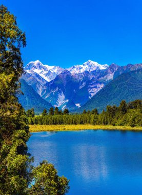 Gorgeous snow-capped mountains are reflected in the smooth cold water of Lake Matheson. South Island of New Zealand. Mount Cook and Mount Tasman. The concept of active and photo tourism clipart