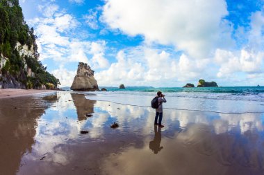 Yeni Zelanda 'ya git. Katedral Koyu 'nda okyanus gelgiti. Kadın okyanusun fotoğrafını çekiyor. Islak kumdaki bulutların ayna yansımaları. Egzotik, ekolojik ve fotoğraf turizmi kavramı