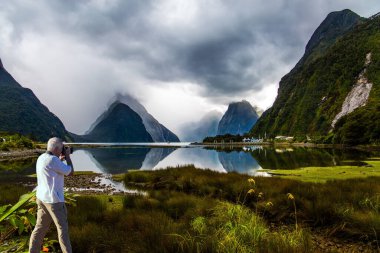 Yeni Zelanda. Meşhur okyanus fiyortu Milford Sound. Gri saçlı turist resimli bir manzara çiziyor. Turist limanı, eğlence gemileri, yatlar ve tekneler. Etkin ve fotografik turnuva kavramı