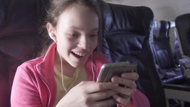 Веселая девочка-подросток играет в игру на смартфоне в салоне самолета во время путешествия на складе видео — стоковое видео