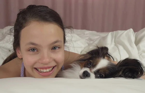 Ευτυχισμένος κορίτσι φιλιά και παίζει με το σκυλί Papillon στο κρεβάτι — Φωτογραφία Αρχείου