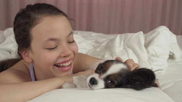 Feliz adolescente menina beijos e brinca com o cão Papillon na cama — Fotografia de Stock