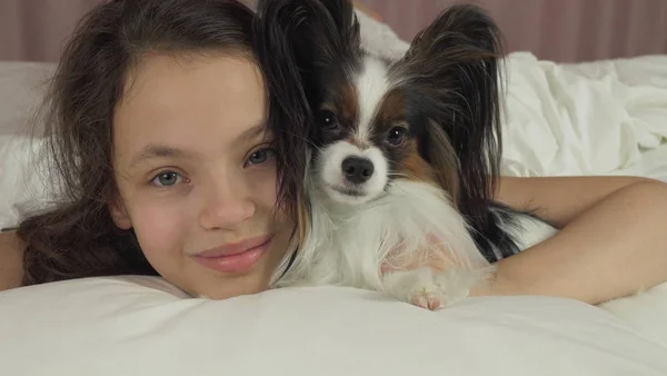 Happy teen girl kisses and plays with dog Papillon in bed — Stock Photo, Image