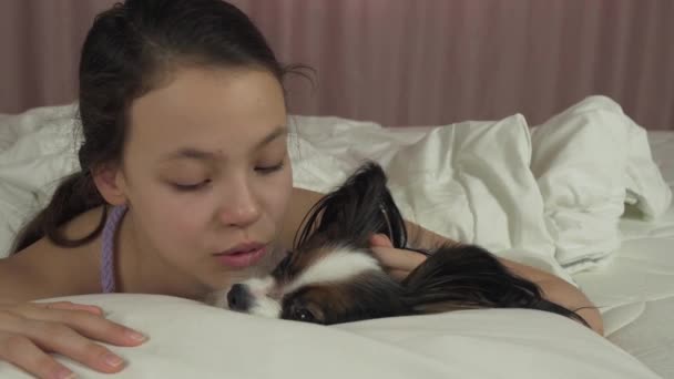 Lycklig tonåring flicka kyssar och leker med hunden Papillon i säng arkivfilmer video — Stockvideo