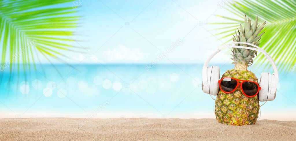 Summer tropical sea with sparkling waves, pineapple with sunglasses and headphones on hot sand beach. Travel and vacation concept with copy space
