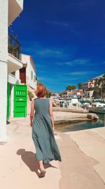 Uzun yaz elbiseli bir kadın, İspanya 'nın Balearic Adaları yakınlarındaki Santanyi, Mallorca yakınlarındaki Cala Figuera adlı büyüleyici balıkçı köyünde yürüyor. Geleneksel Akdeniz tarzı kıyı evleri yakınlarında demirlemiş küçük tekneler.