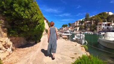 Uzun yaz elbiseli bir kadın, İspanya 'nın Balearic Adaları yakınlarındaki Santanyi, Mallorca yakınlarındaki Cala Figuera adlı büyüleyici balıkçı köyünde yürüyor. Geleneksel Akdeniz tarzı kıyı evleri yakınlarında demirlemiş küçük tekneler.