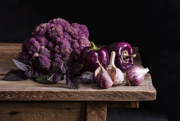 Fresh vegetable. Dark purple peppers, cauliflower with leaves of basil and garlic on old rustic wooden table on black background