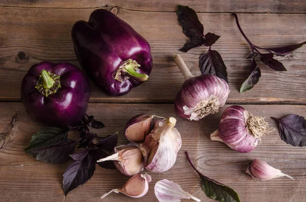 Dark purple peppers with leaves of basil and garlic on old rustic wooden table on black background. Top view