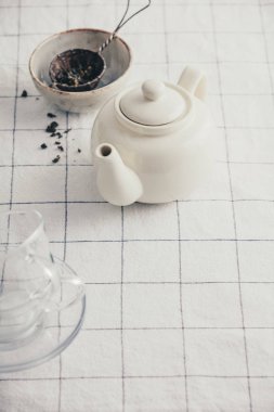 Kareli masa örtüsünde beyaz çaydanlık, bardak ve süzgeç.