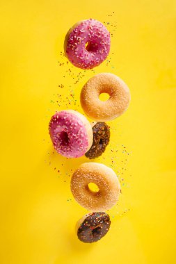 Çeşitli dekore edilmiş donutlar, hareket halinde serpiştirilmiş, sarımsı arka plana dökülüyor.