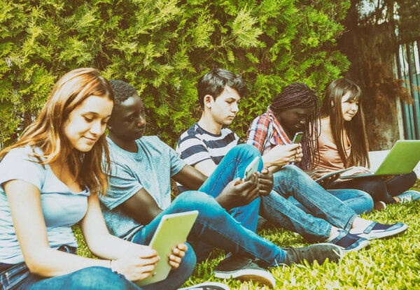 Teenagers of different races using tablets, smartphones and laptops in a city park.