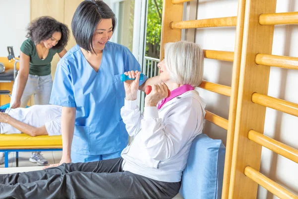 Yaşlı kadın küçük ağırlık içinde rehabilitasyon dacility, öğretmenler tarafından yardım ile.