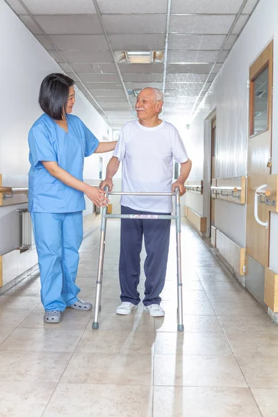 Asian doctor helping elder man with walker in hospital hallway.