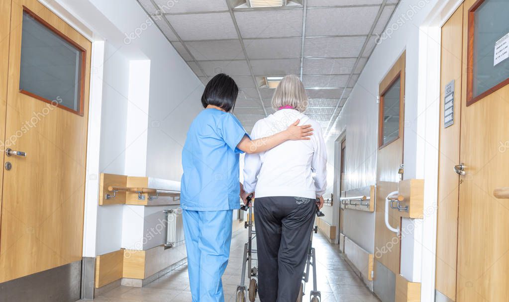 Asian doctor helping elder woman with walker in hospital hallway.