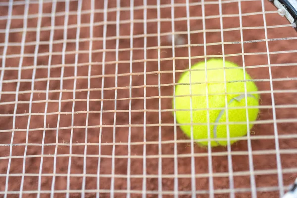 Yellow tennis ball behind racket cords.