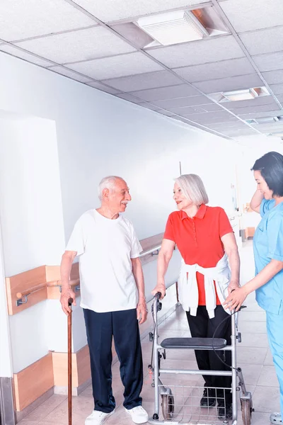 在护士的协助下 在医院走廊里移动棍子和步行者的老年夫妇 — 图库照片