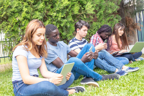 Teenagers outdoor using laptop and tablet seated on the grass.