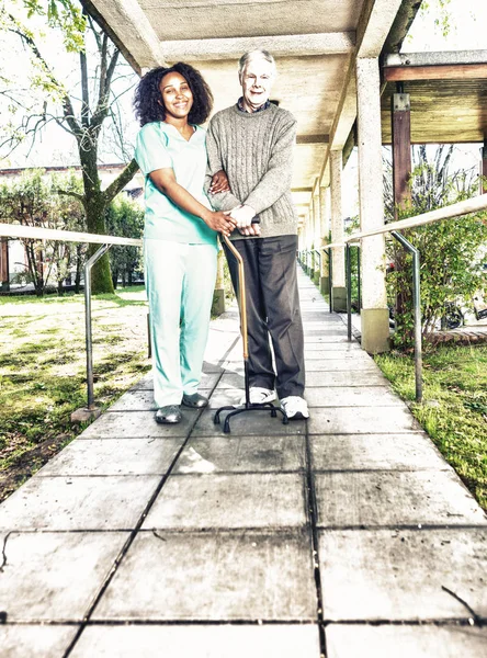 非洲护士帮助老年男子在康复设施走廊的拐杖 — 图库照片