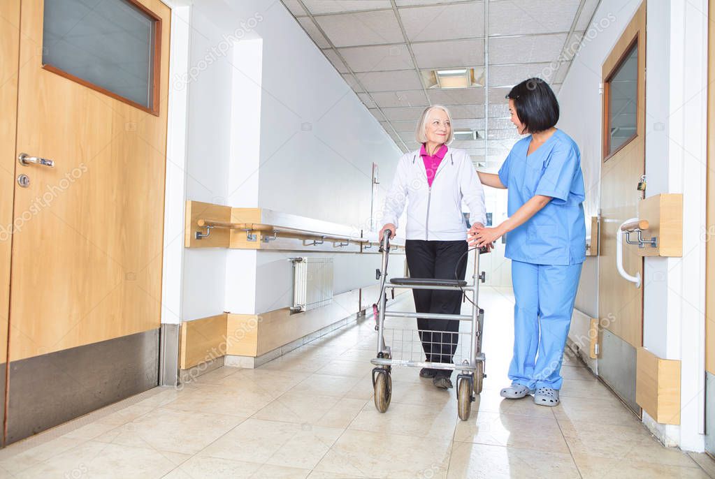 Asian nurse assisting aged senior woman with walker in rehab fac