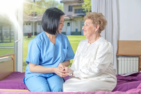 亚洲女医生牵着她的手 与坐在病床上的成熟老年妇女嬉笑着玩耍 退休社区概念 — 图库照片