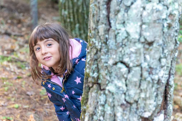 Chica joven jugando a las escondidas detrás de un tronco de árbol — Foto de Stock