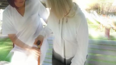 Genç Asyalı kadın doktor hastane bahçesindeki bankta oturan yaşlı kadına yardım ediyor. Arkadan gelen ışık ışınları