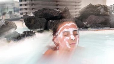 Genç bir kız sıcak bir termal banyoda yüzüne krem sürüyor. Ağır çekim. Gökyüzünden gelen güneş ışınları