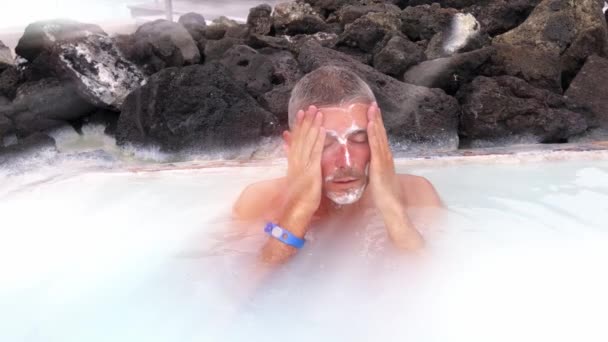 Человек, намазывающий лицо кремом в горячей термальной ванне. Медленное движение. Солнечные лучи, исходящие с неба — стоковое видео