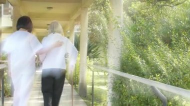 Genç Asyalı kadın doktor hastane bahçesinde koltuk değnekleriyle yaşlı kadına yardım ediyor. Arkadan bak. Arkadan gelen ışık ışınları