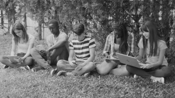 Gruppo di razze miste adolescenti seduti sull'erba che giocano con gadget tecnologici, filtro in bianco e nero — Video Stock
