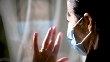 Ağız maskesi takan kadın Coronavirus saatinde pencereden dışarı bakıyor.