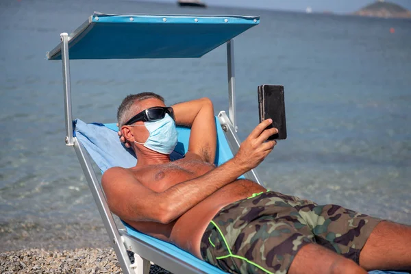 Biały Mężczyzna Niebieskiej Masce Chirurgicznej Siedzący Leżaku Plaży Czytający Booka — Zdjęcie stockowe