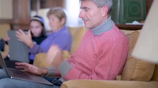 Een thuiswerkende man, zittend op de bank met laptop, zijn moeder en dochter in dezelfde kamer. Langzame beweging — Stockvideo