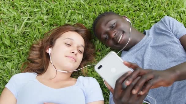 Wieloetniczna para nastolatków siedząca w parku słuchając muzyki. Zwolniony ruch — Wideo stockowe
