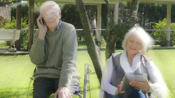 Couple retraité jouant avec la tablette et parlant au téléphone portable en plein air. Rayons lumineux par derrière — Video