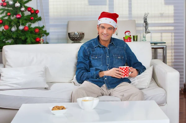 自宅でクリスマスシーン 40代の男は帽子をかぶってソファの上でリラックスしてコーヒーを飲んで — ストック写真