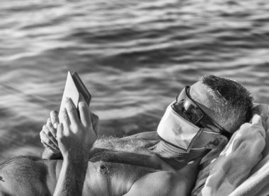 Maske takan ve sahilde tablet okuyan bir adam..