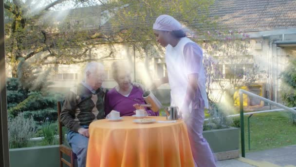 Медленное движение пожилой пары, завтракающей под открытым небом, обслуживаемой медсестрой в больничном саду. Концепция реабилитации и ухода на пенсию — стоковое видео