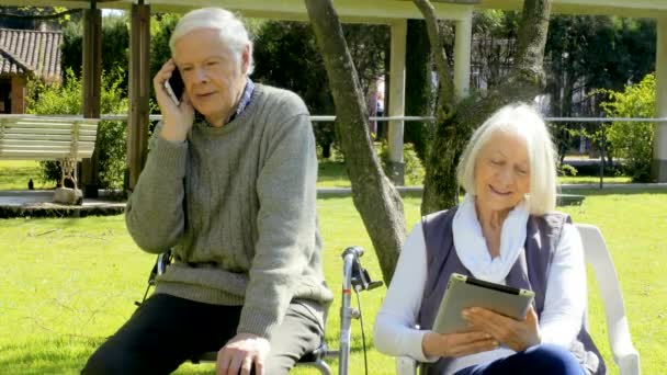 Movimiento lento de la pareja de ancianos jubilados al aire libre en el jardín del hospital de rehabilitación con teléfono inteligente y tableta. Concepto de felicidad, rehabilitación y jubilación — Vídeos de Stock