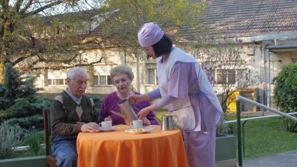 Slow motion of elderly retired couple having breakfast outdoor served by nurse in hospital garden. Rehabilitation and retirement concept — Stock Video