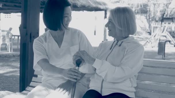 Anziana donna seduto su il panchina aiutato da asiatico femmina infermiera — Video Stock