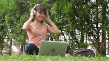 Parkta dizüstü bilgisayarıyla müzik dinleyen mutlu Asyalı kız. Yavaş çekim.