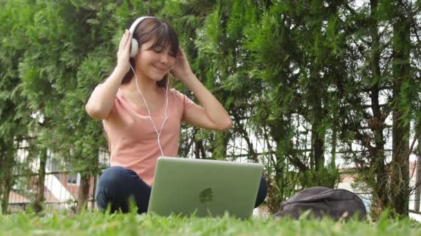 Šťastná asijská dívka slyší hudbu v parku se svým laptopem. Zpomalený pohyb. — Stock video