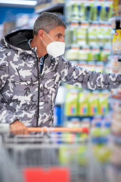 在一场大脑皮层病毒大流行期间 戴着防护面罩 提着购物车的男人站在食品杂货店里挑选食物 — 图库照片