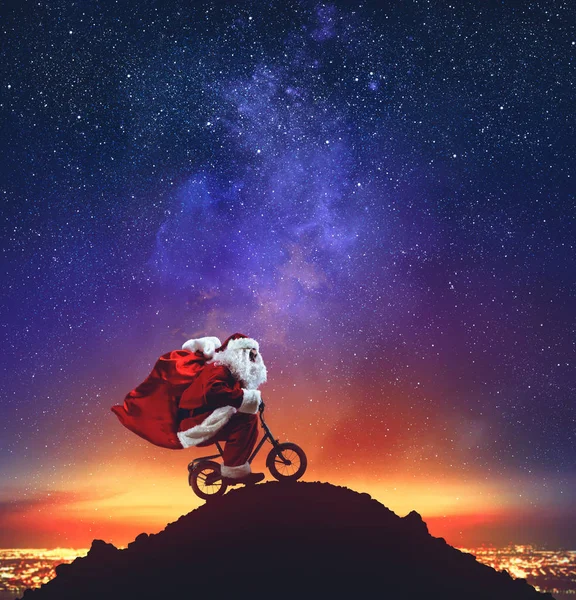 Santa Claus on a little bike on the peak of a mountain under the stars — Stock Photo, Image